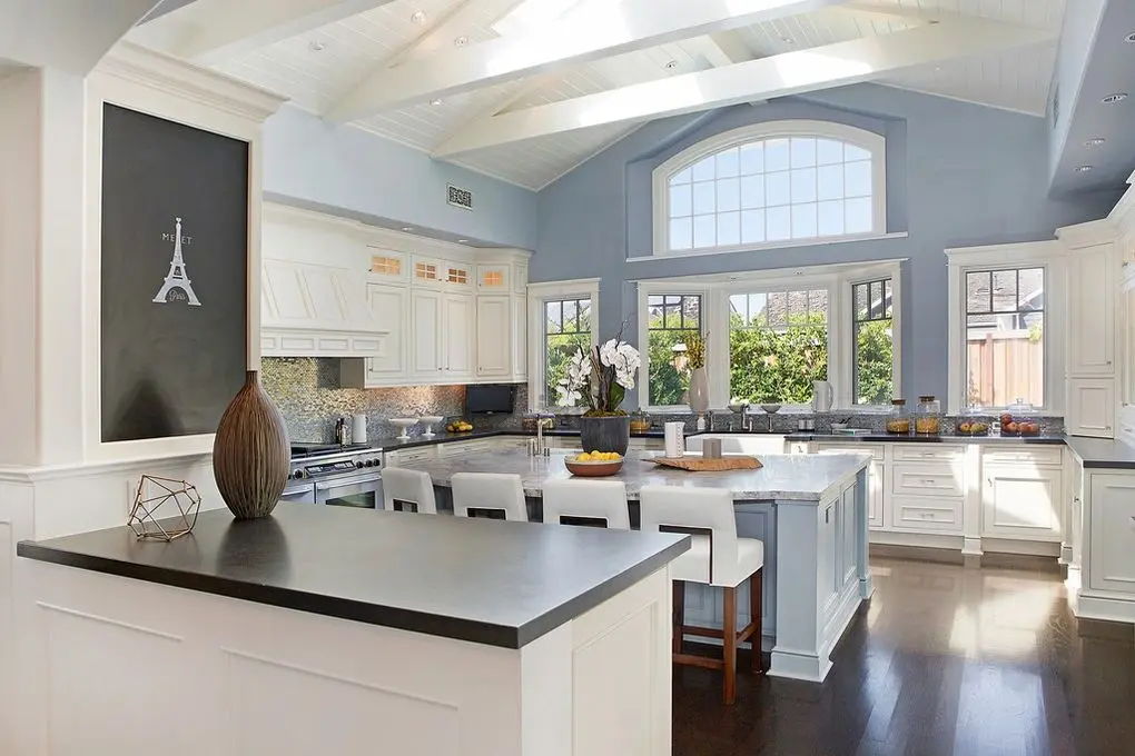 Bright kitchen in the 'Port Streets Remodel' with white cabinetry, marble countertops, and a central island, all under a vaulted ceiling with skylights.