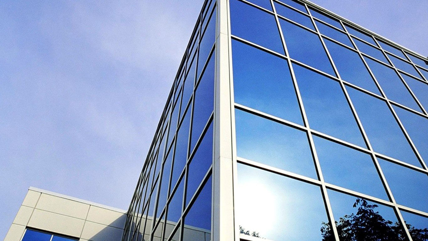 La façade en verre du bâtiment reflète un ciel bleu et une partie du soleil, créant ainsi un aspect moderne et élégant. Les silhouettes des arbres sont visibles dans le reflet inférieur.