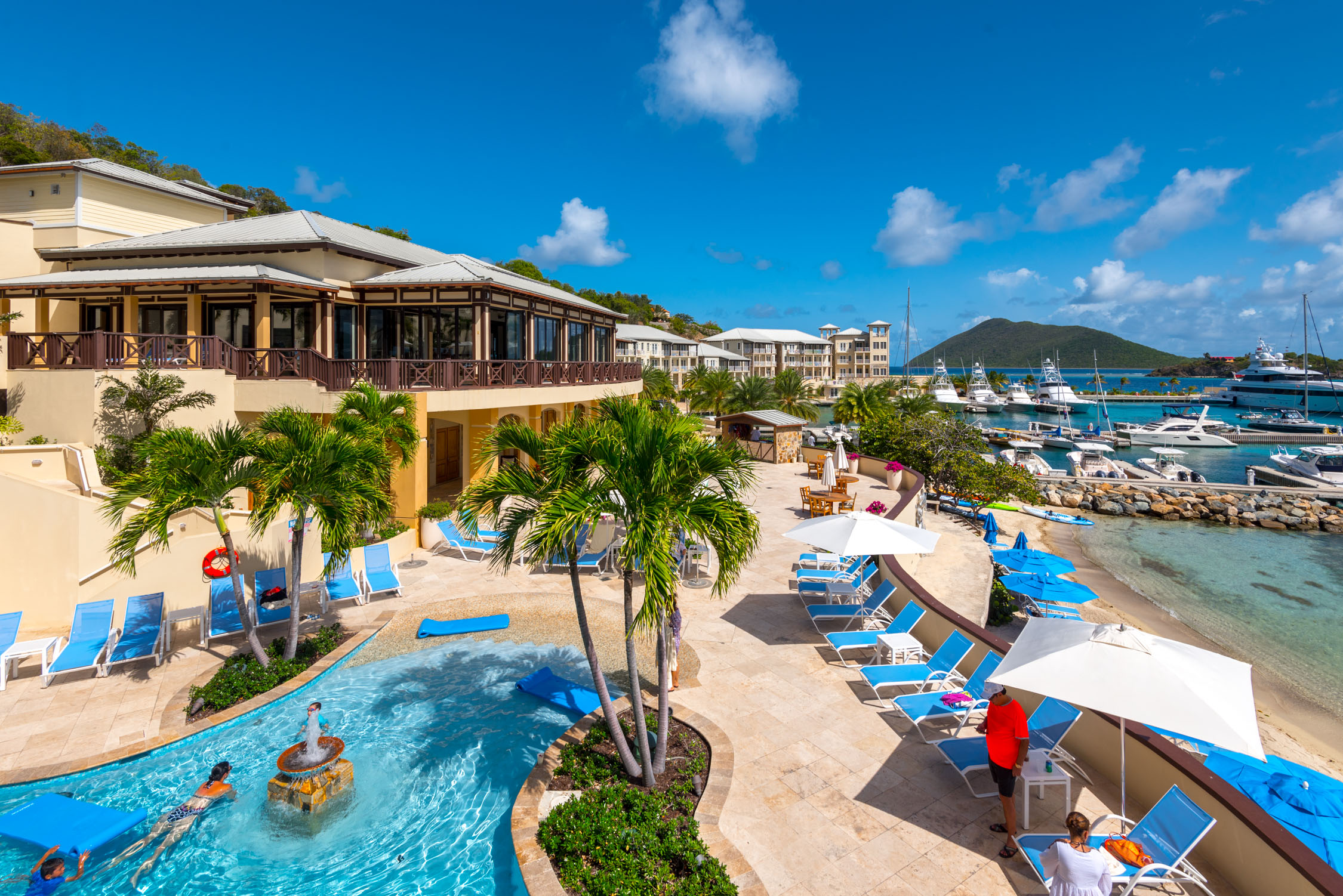 A tropical beach resort with a swimming pool, lounge chairs under umbrellas, and a restaurant overlooking turquoise water.