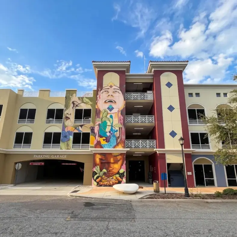 The image features a vibrant mural on a parking garage in Bradenton, FL, showcasing colorful, artistic designs against a backdrop of a clear blue sky with scattered clouds.