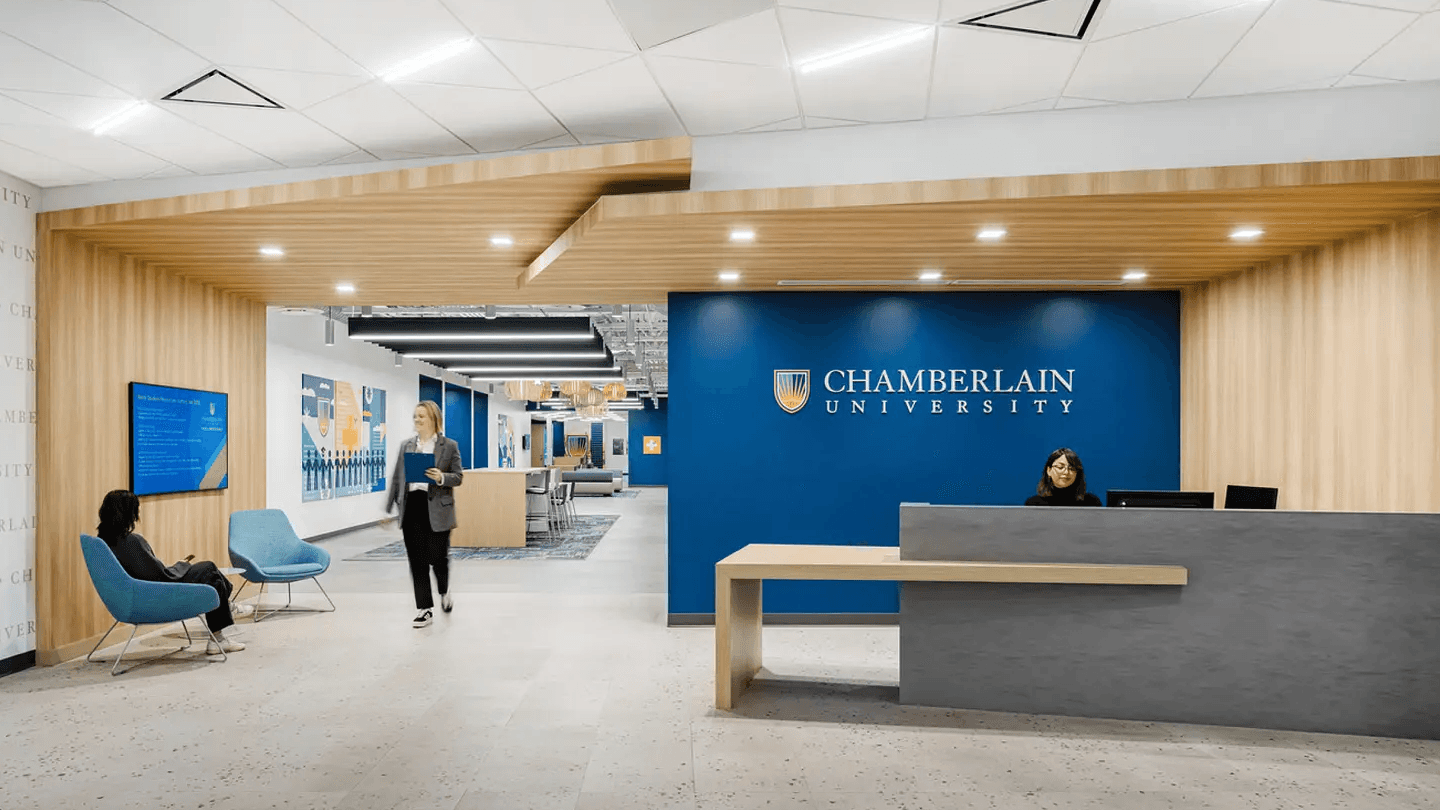 Modern university lobby with a blue feature wall displaying "Chamberlain University" logo, a reception desk, and seating. Bright, welcoming atmosphere.