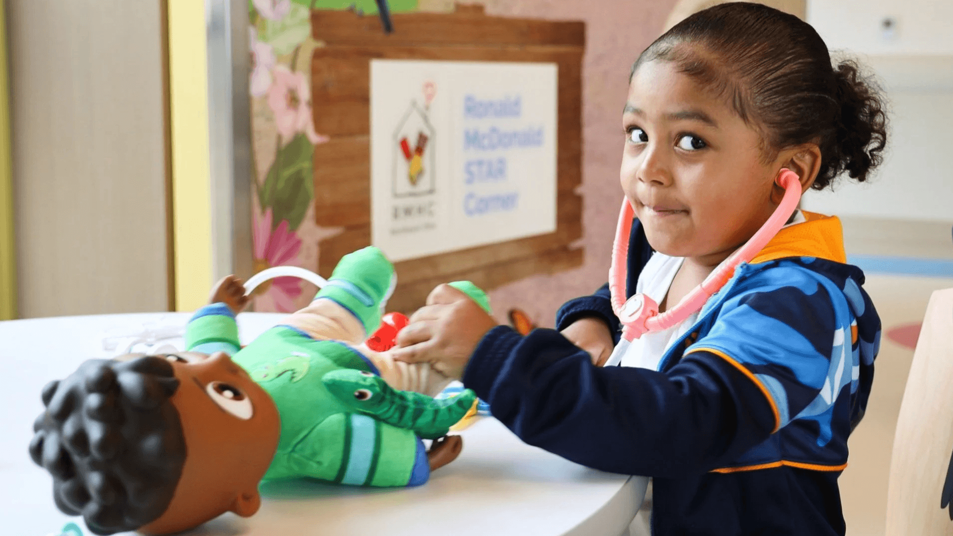 Young child using a toy stethoscope to examine a doll during pretend medical play at a table in a colorful indoor setting.