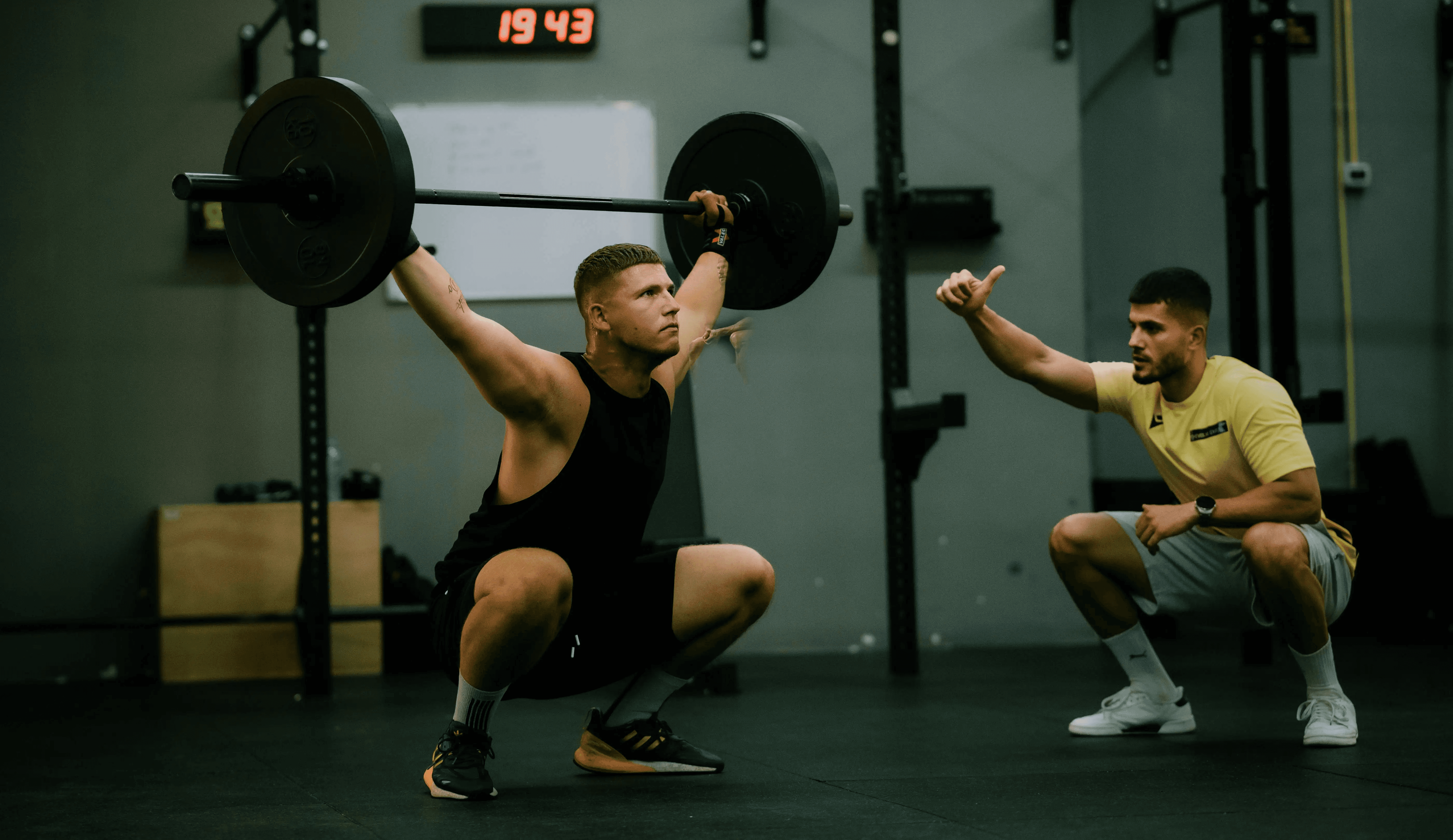 Weightlifter performing an overhead squat under supervision in a gym.