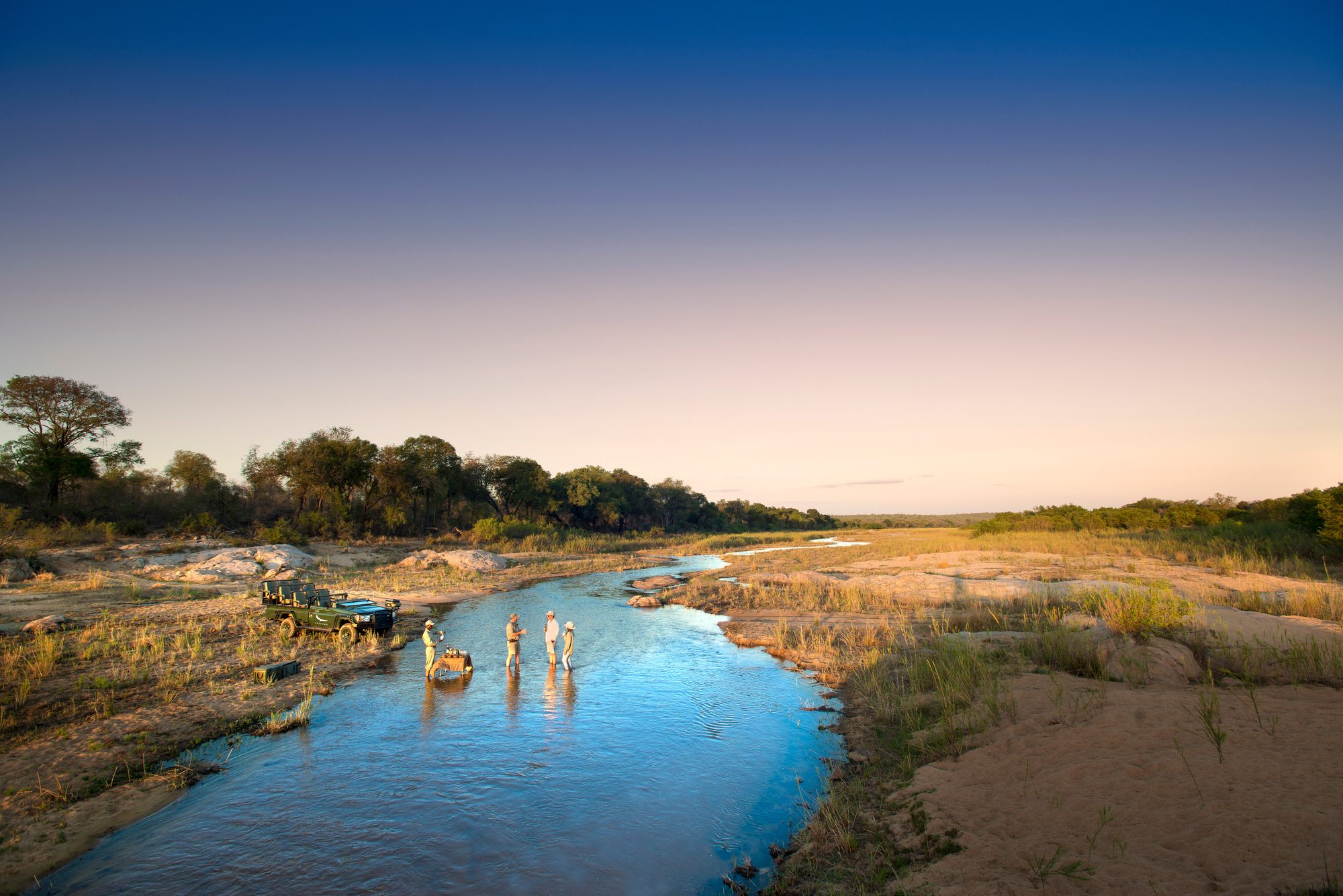 Two guides and two guests gather with drinks around a small table set up in a shallow river at sunset in South Africa, with an open safari vehicle parked on the sandy bank.