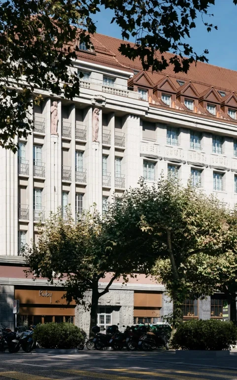 A large, elegant building with a red-tiled roof and tall columns stands behind leafy trees and parked scooters; a white van is parked on the street in front under a clear blue sky.