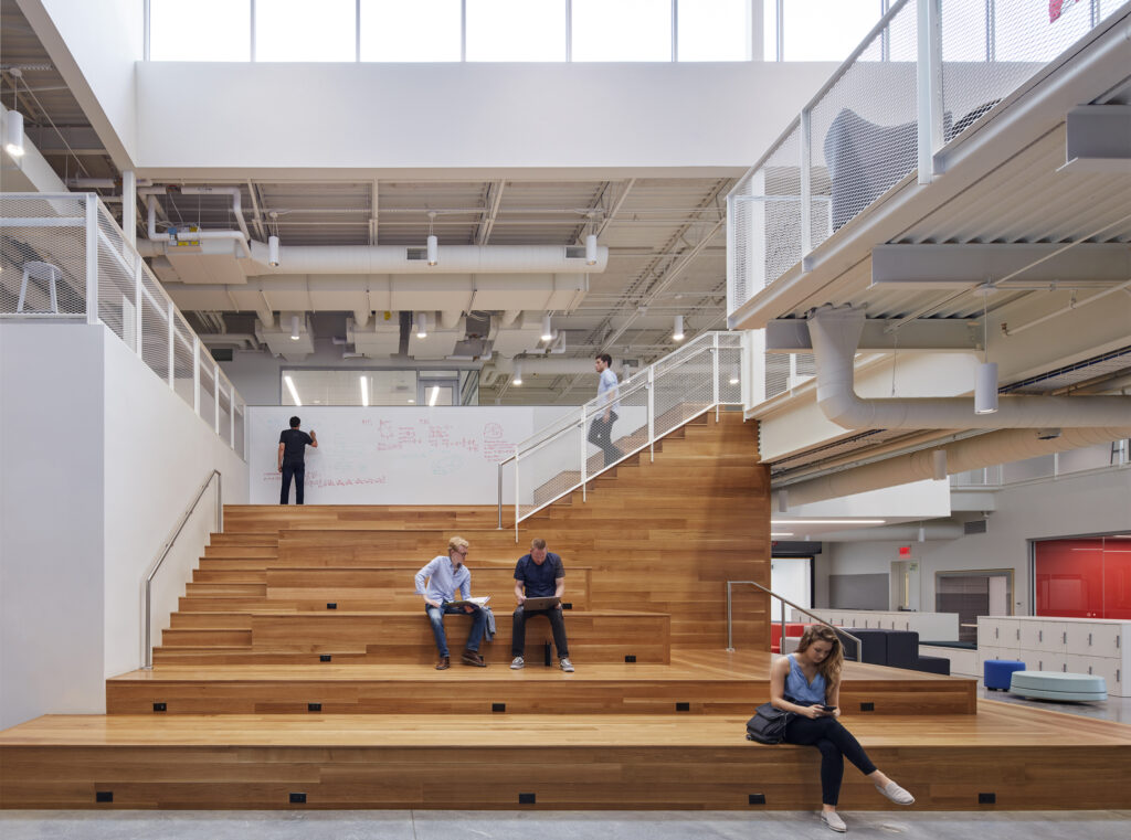 Interior of BVH Architecture office featuring a wide wooden stadium-style seating staircase. Several people are using the space casually - some collaborating at a whiteboard, others sitting and working on the steps. The space has an industrial-modern aesthetic with exposed ceiling systems and white walls.