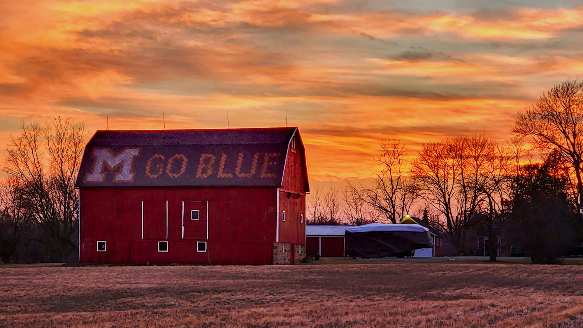 A red barn stands in a field under a dramatic orange and purple sunset, with trees silhouetted in the background.