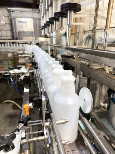 Green plastic bottles on a conveyor belt in a chemical manufacturing facility, being filled by automated machinery.