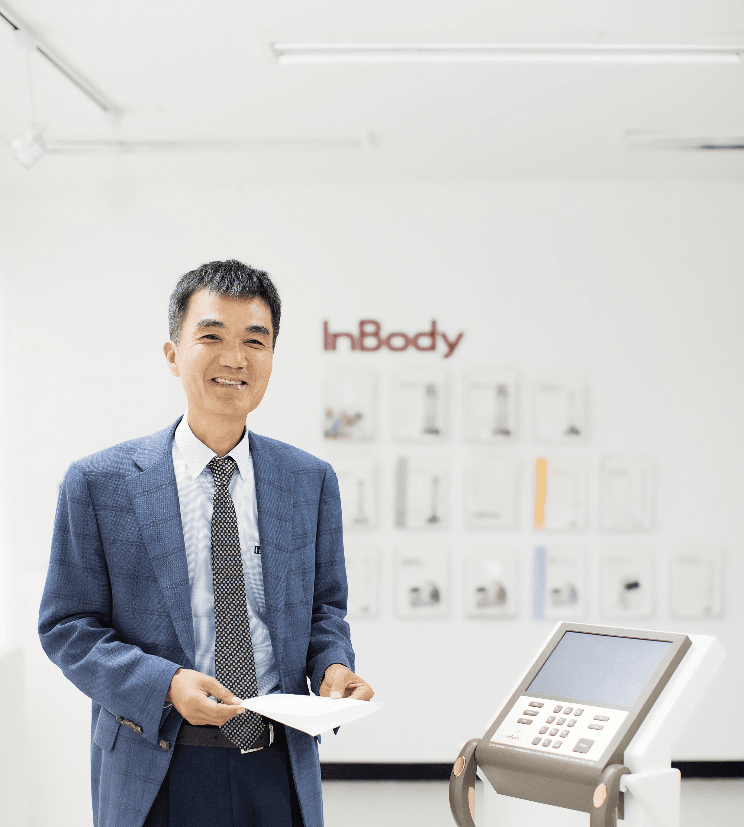 InBody CEO Dr. Cha Ki-chul wearing a blue plaid suit and smiling while standing next to an InBody body composition analyzer, with the company logo visible on the wall behind him.