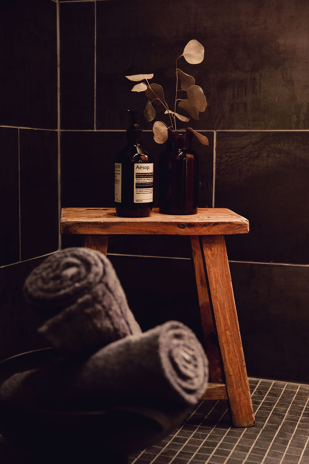 Aesop soap and dried eucalyptus sit on a wooden bench inside the shower in the greenroom of Particle Studios in Seattle, WA. Two towels are neatly rolled and displayed in the foreground.