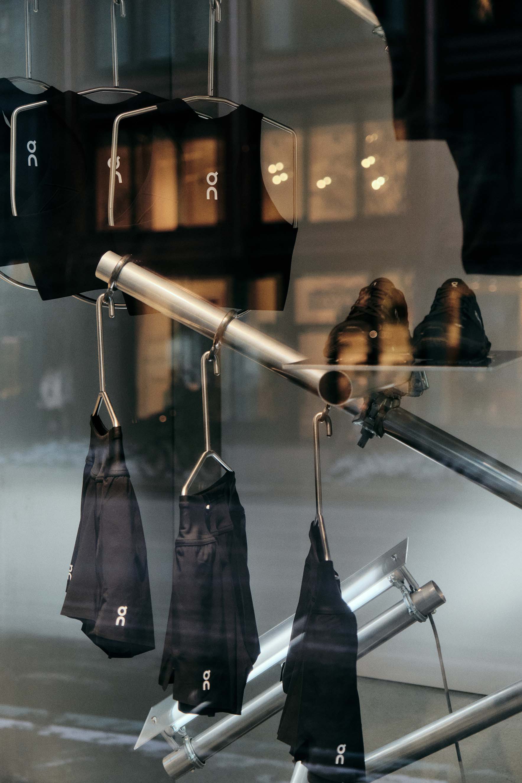 A store display features black athletic shoes, shorts, and shirts with the On brand logo, all neatly hung on silver metal rods—a striking campaign that captures city street reflections in the glass.