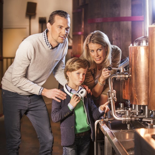 Egy háromfős család gépeket vizsgál egy teremben, ahol a fiú mutat és fejhallgatót tart a kezében, miközben a szülők figyelik.