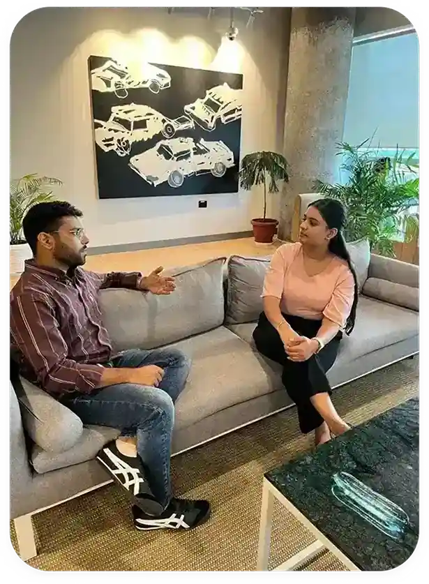 A young man and woman engage in a serious conversation while seated on a light gray couch in a modern, well-lit office environment, featuring potted plants and abstract car artwork on the walls, representing a setting for Unbiased Counselling related to UG/PG Online Degree Programs with EazyGrad.