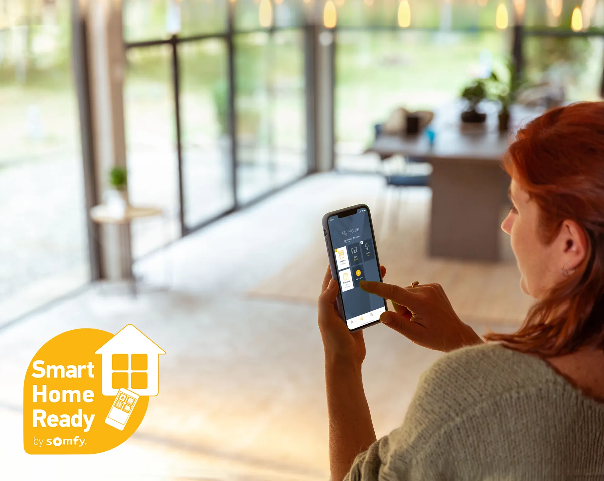 Frau bedient Smartphone mit Somfy Smart Home App im hellen Wohnzimmer, Logopräsenz.