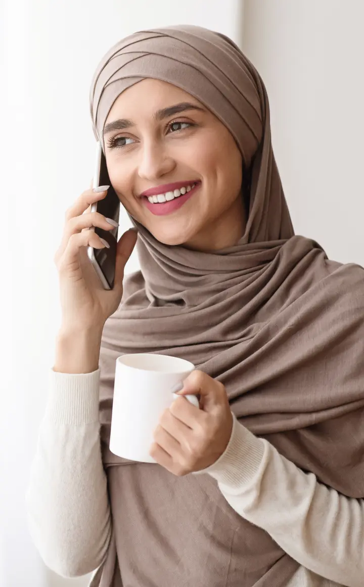 A smiling woman in a hijab holding a coffee cup while talking on the phone, representing a relaxed and friendly interaction, possibly related to health coaching or support.