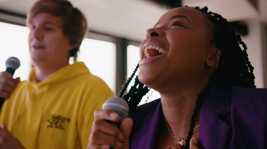 A young person with curly hair smiles at the camera while sitting in front of a laptop against a dark background.