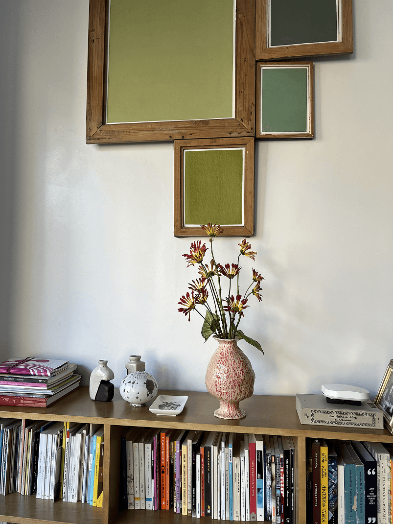 Jarrón rosado con flores rojas y amarillas sobre un estante de madera con libros y objetos decorativos, frente a una pared con marcos de colores verdes.