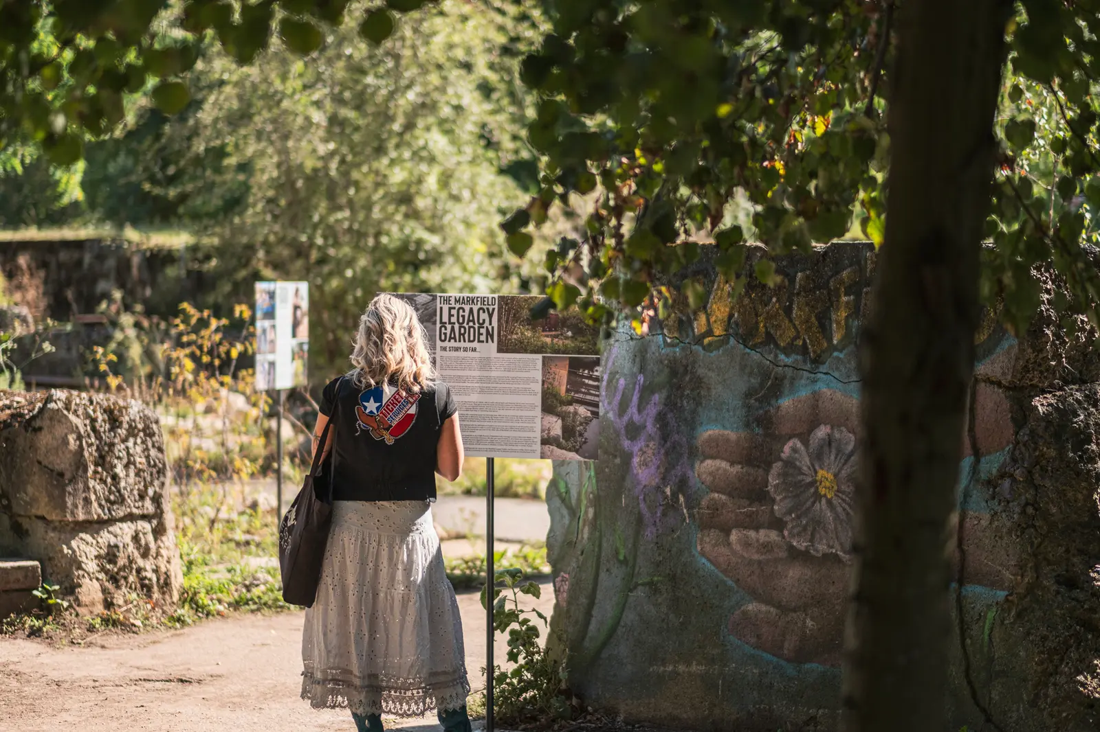 A woman walks along a path surrounded by greenery, with a textured wall in the background. The scene is sunny.