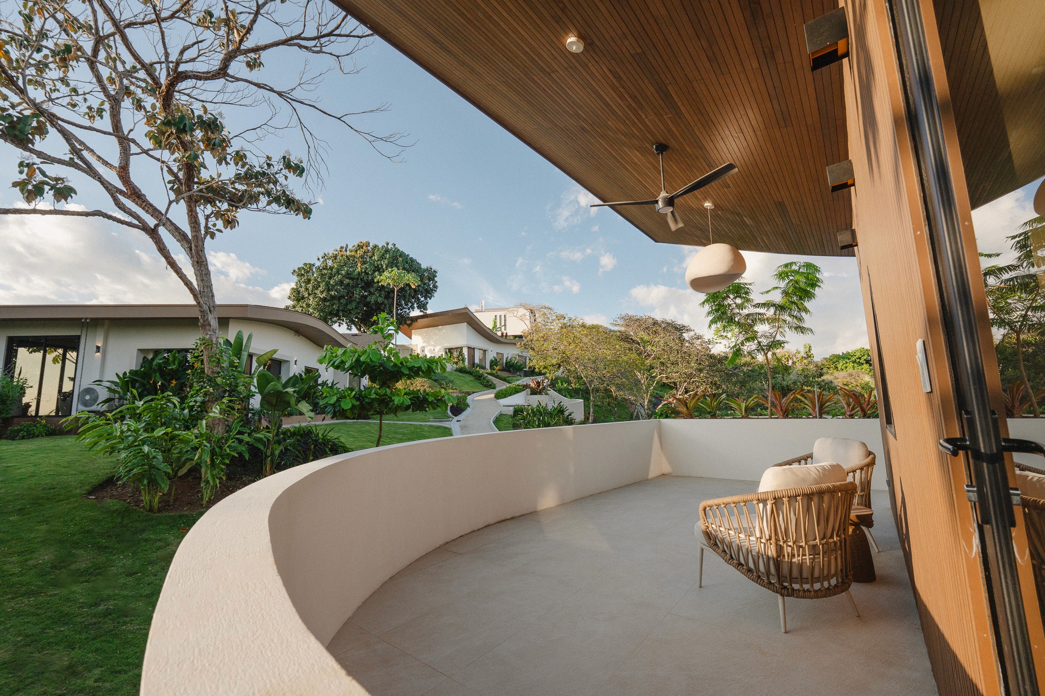 Private curved balcony of a luxury vacation rental in Costa Rica. Features include two woven lounge chairs, a wood-paneled ceiling with a fan, and a panoramic view of the tropical gardens and surrounding modern architecture.