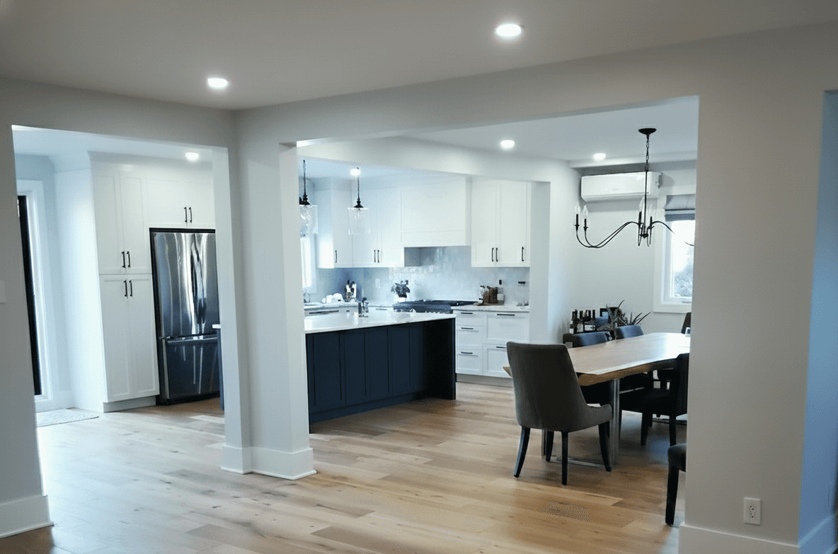 L'îlot cetral est bleu foncé. Le comptoir de la cuisine est en quartz blanc. Les armoires sont blanches, style shaker. Les poignées sont noires et fines. Le réfrigérateur est en acier inoxydable et encastré. La cuisinière est professionnelle sous une hotte blanche décorative. Deux suspensions en verre pendent au-dessus de l'îlot. Des spots encastrés éclairent toute la cuisine et les pièces. La table à manger est en bois brun massif. Les chaises sont grises anthracite, rembourrées avec dossier haut. Un lustre noir de style classique surplombe la table. de larges ouvertures séparent les espace. Des piliers et poutres blanches sont apparents. Le sol est en bois clair. Les murs sont gris très pâle. L,ensemble est lumineux et aéré.