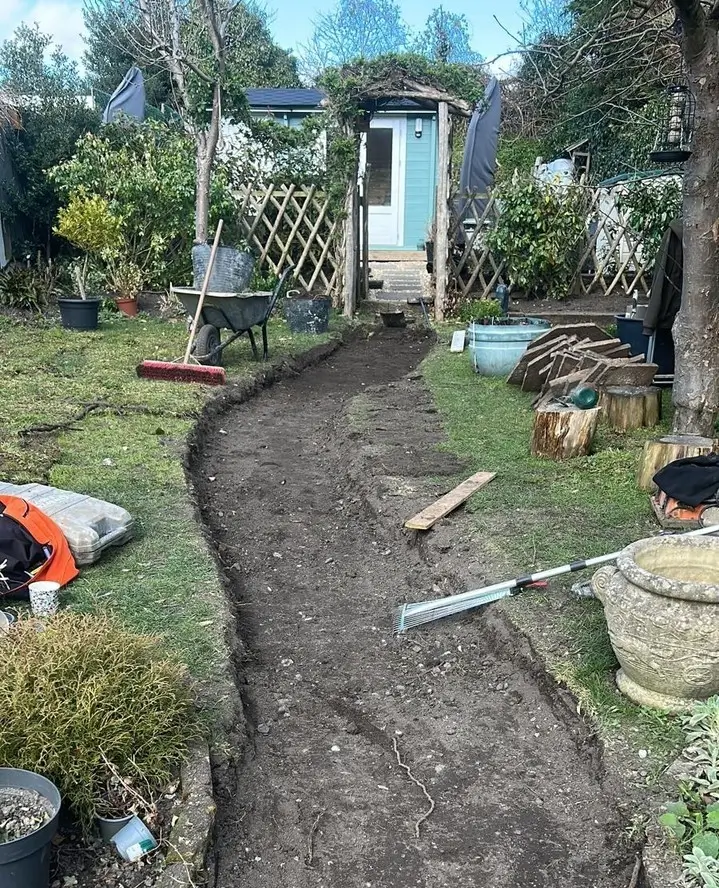 A garden path under construction, surrounded by greenery and tools, leading toward a shed in the background.