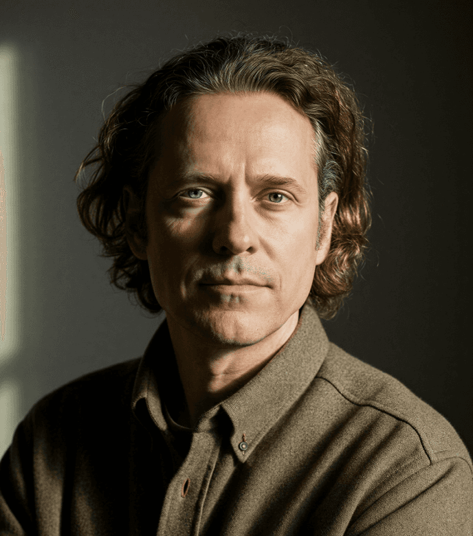 Portrait of a man with curly shoulder-length brown hair wearing a brown flannel button-up shirt, looking directly at the camera with a calm expression against a dark background with dramatic side window light.