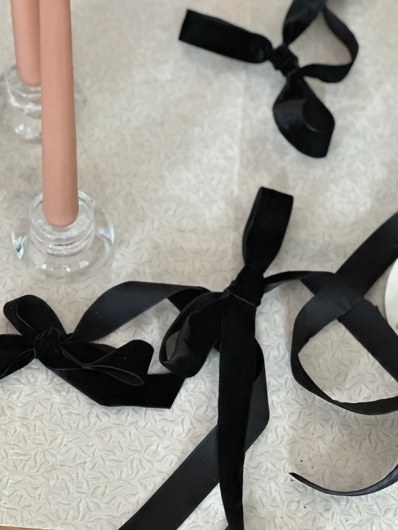 Lazos de terciopelo negro dispuestos sobre un mantel claro con textura, junto a candelabros de vidrio con velas rosas.
