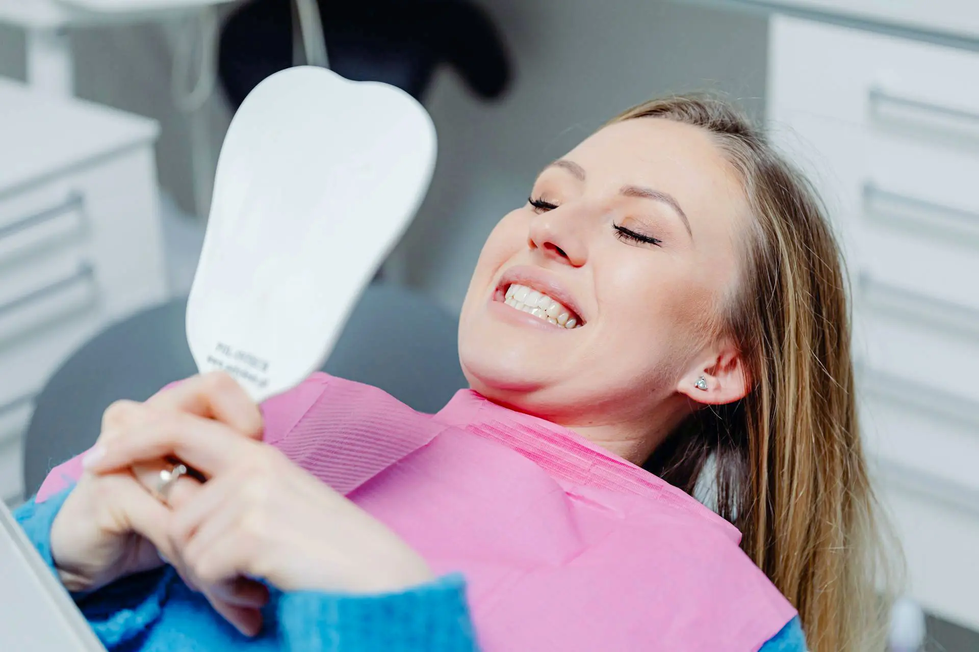 A smiling woman in a dental chair holds a mirror, wearing a pink bib and looking at her teeth.