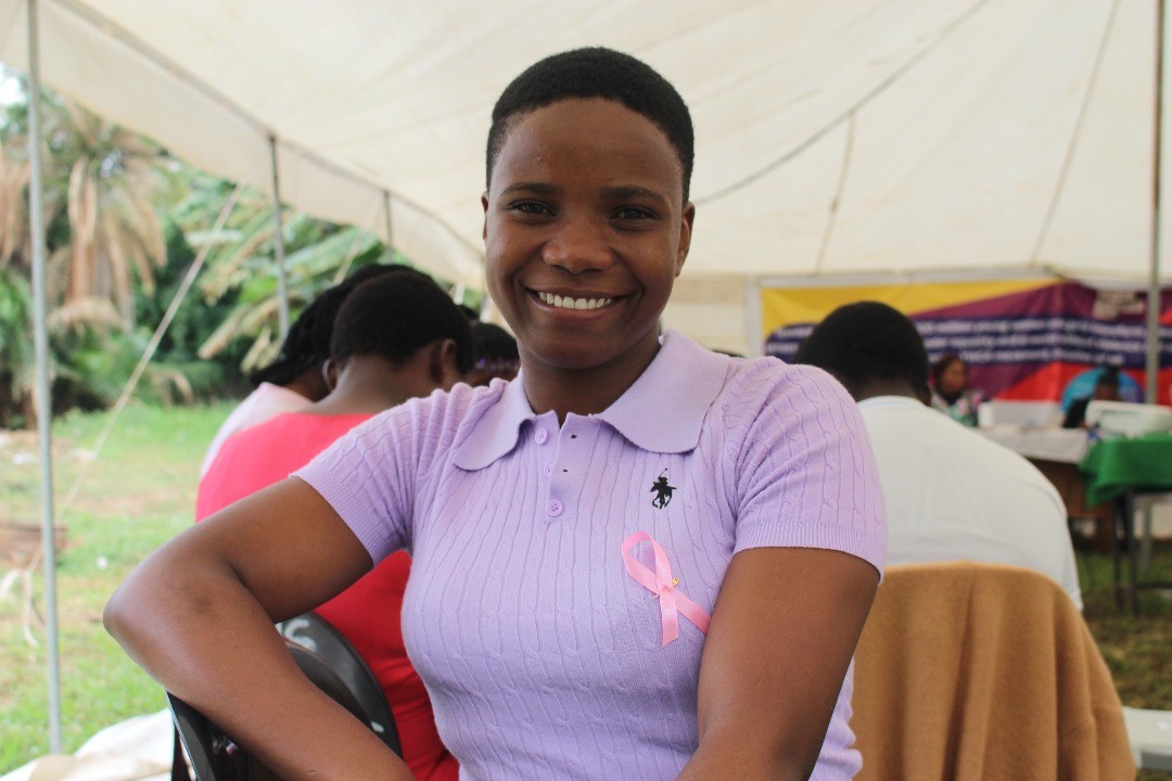 A smiling person with curly hair sits at a desk, looking at a laptop against a dark background.