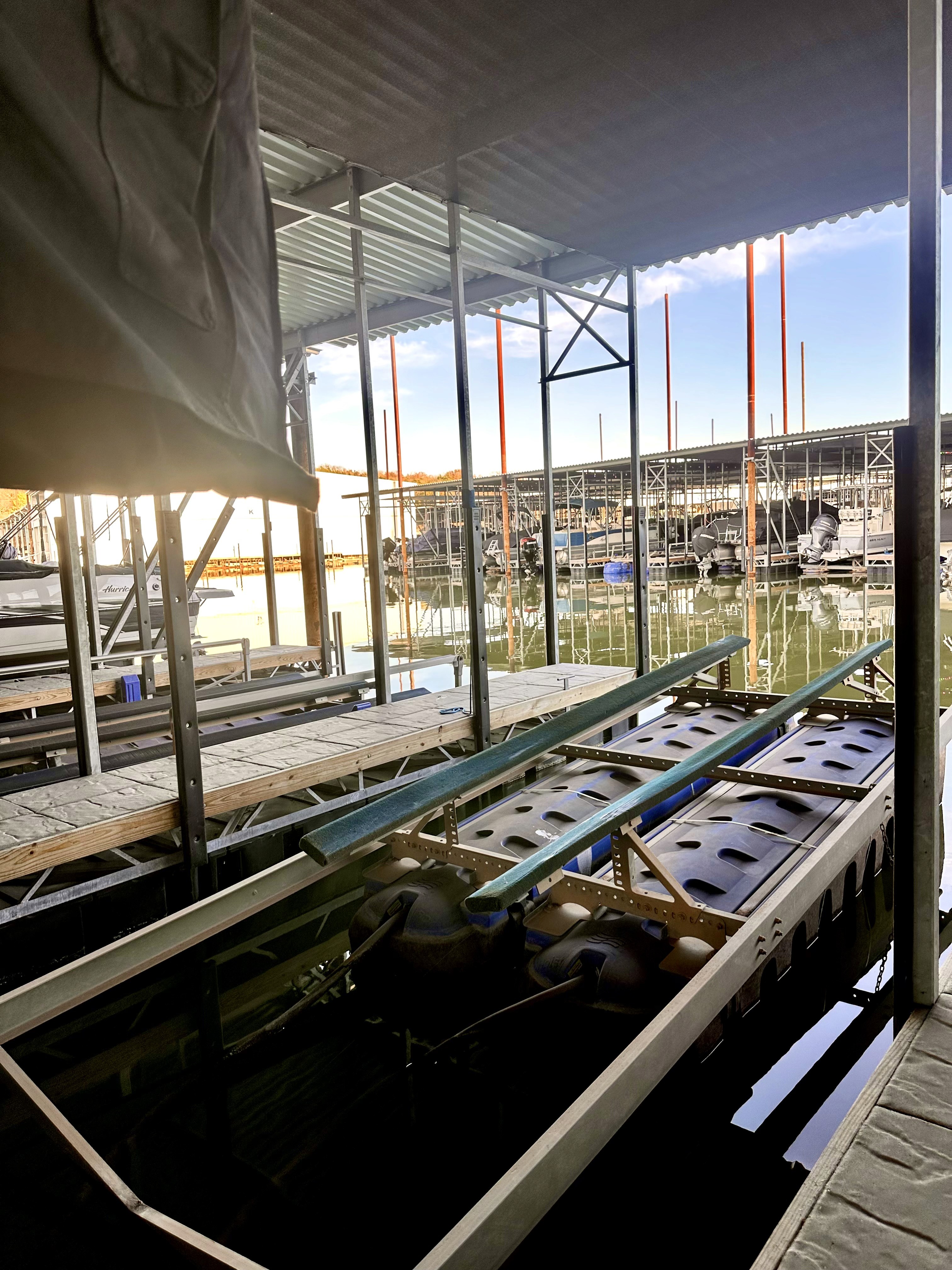 A docked boat rests inside a covered marina, surrounded by calm water and empty slips, with a clear blue sky visible through the open sides of the shelter.