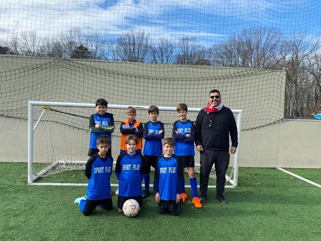 A SportPlus Charlotte competitive soccer team posing with the head soccer coach on a turf field in front of a goal