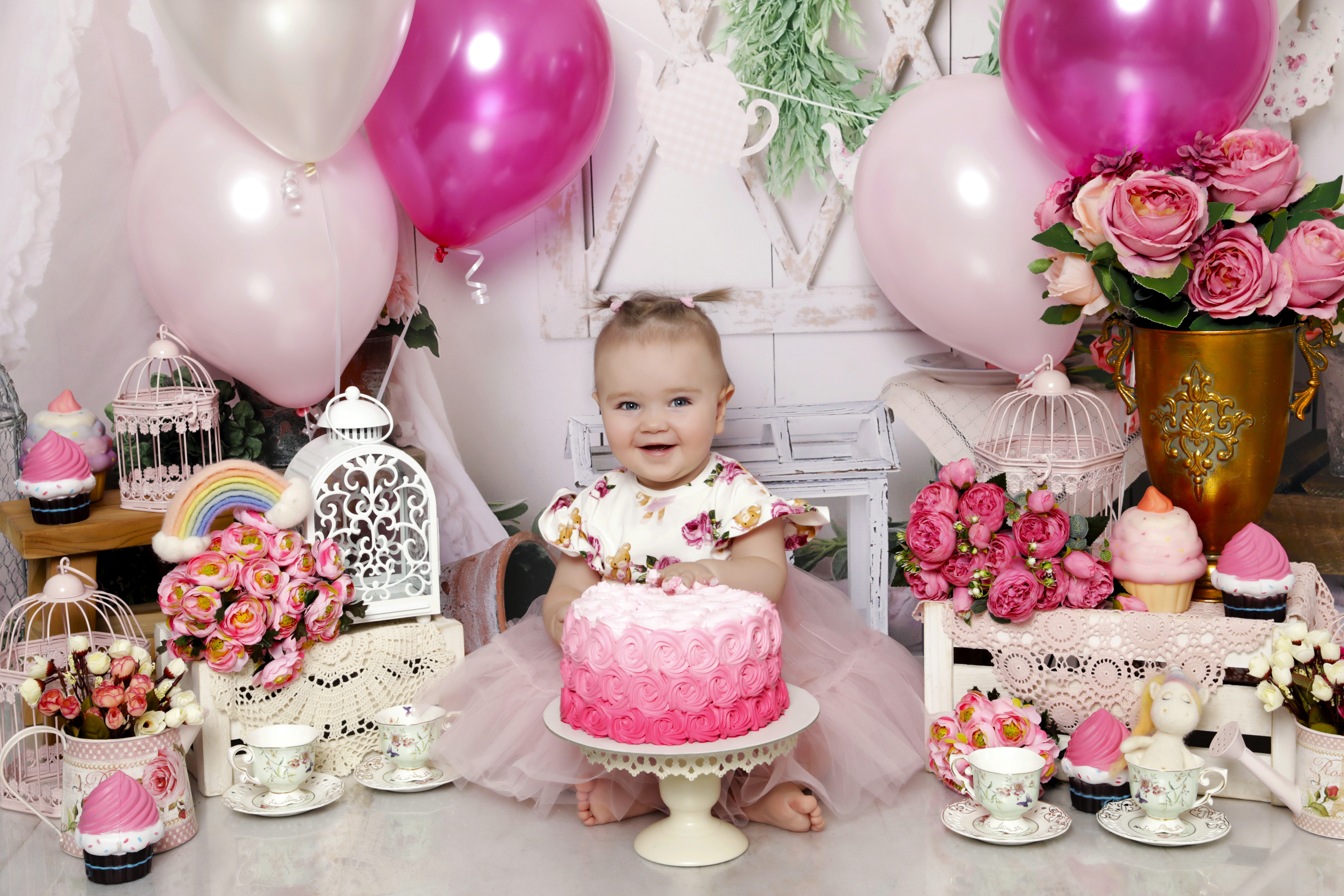 A smiling baby girl is in a pink dress, sitting behind a flowershop-themed photoshoot. There are pink balloons, flowers, and party decorations.