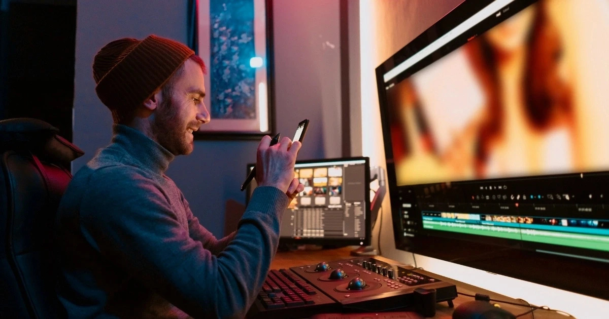 Person smiling while holding a smartphone at a desk with multiple monitors showing video editing software and color grading controls in a dimly lit workspace.