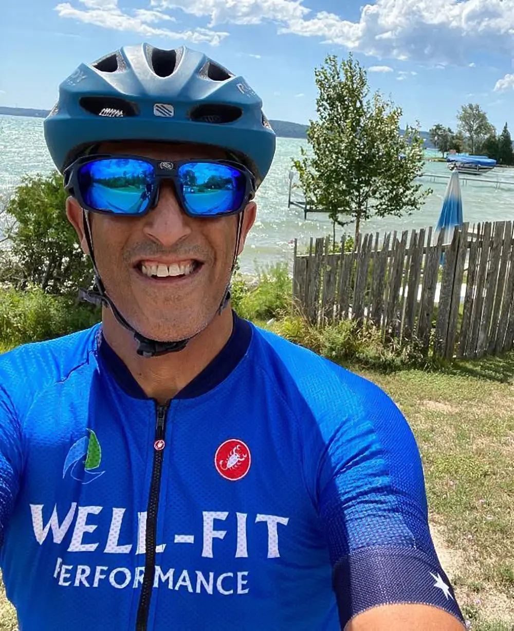 Ironman coach Sharone Aharon smiling in his Well-Fit Performance cycling kit and helmet during a training ride beside a lake