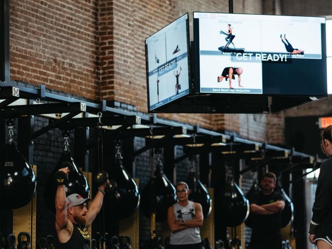 Command center screens at Earn Your Stripes showing the next exercises while a member does a dumbbell press