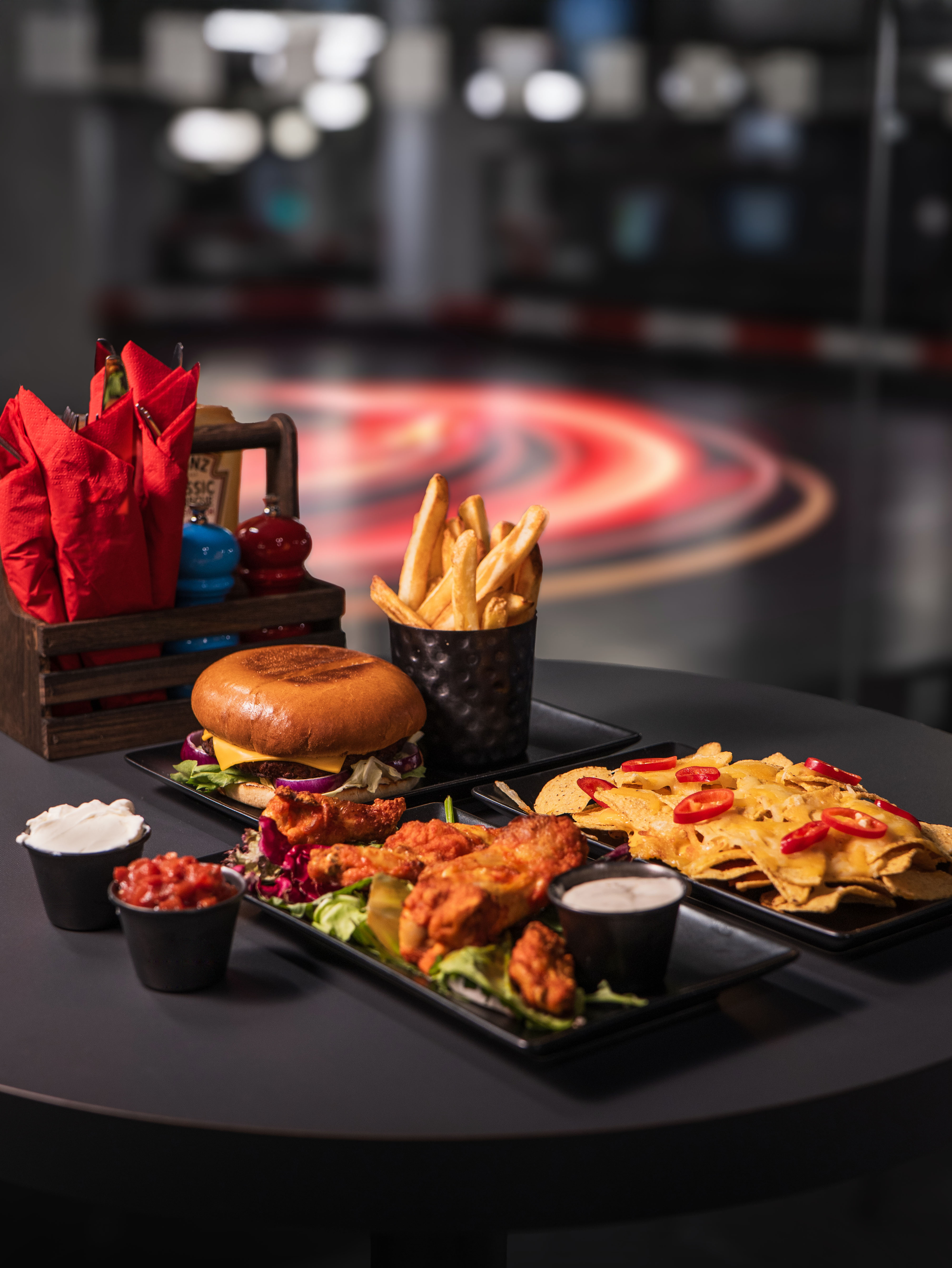 A platter with a burger, fries, chicken wings, sauces, and drinks, set against a dark, modern backdrop.