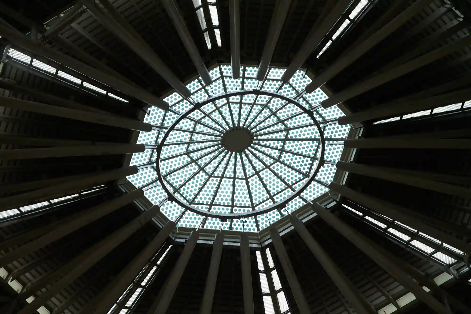 Looking directly up at a large, symmetrical geometric glass skylight supported by dark structural beams in a modern building.