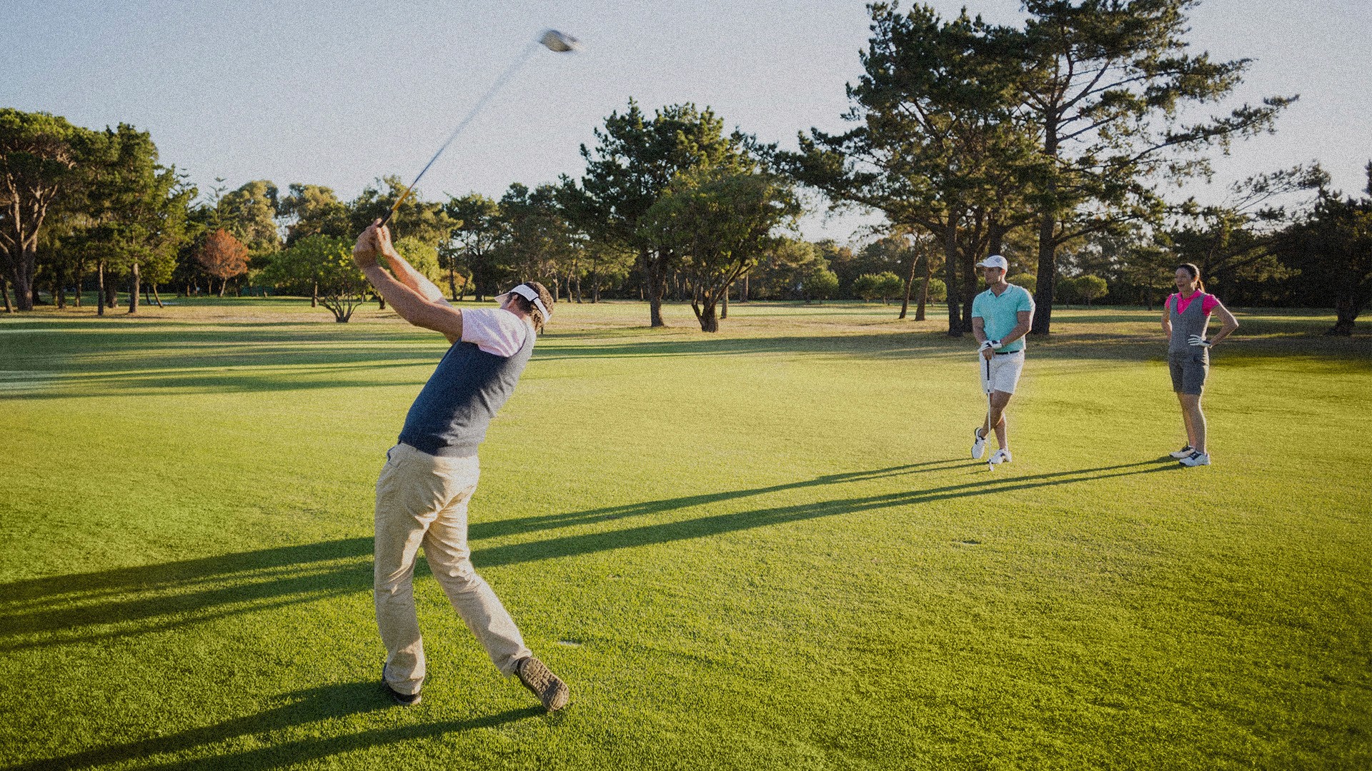 A group of people golfing on a course. one person is swinging while two stand by.