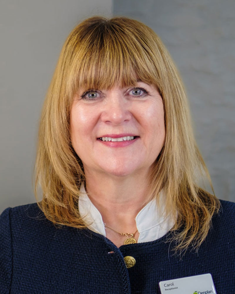 A portrait of Carol, a receptionist at Cricklade Dental Practice, smiling and wearing a white blouse under a dark blue textured jacket with gold buttons. She is wearing a name badge that reads "Carol" and a gold necklace, standing against a light grey textured wall.