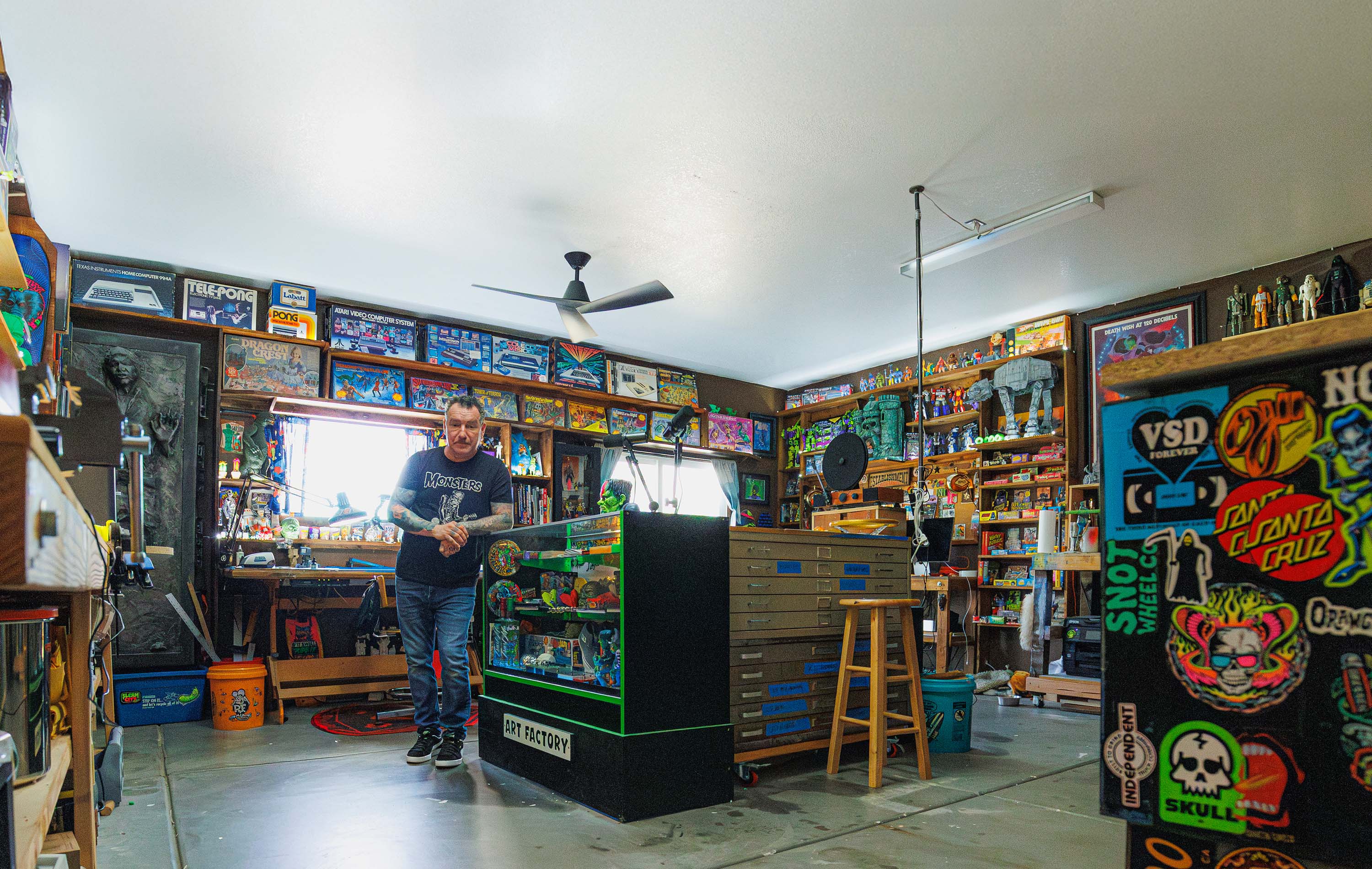 Artist Dirty Donny Gillies standing in his brightly lit studio filled with vintage toys, art supplies, and colorful collectibles, surrounded by shelves packed with models, posters, and pinball machines.