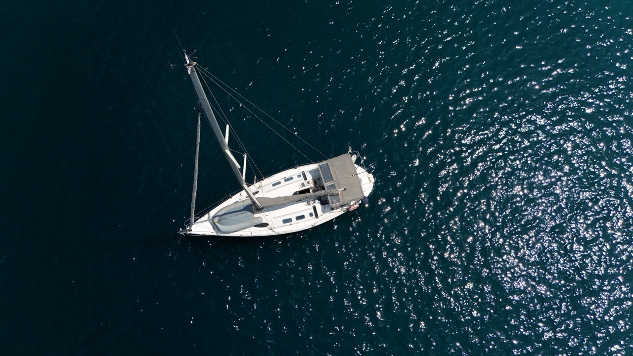 Aerial view looking down on a Beneteau First 38.5 sailing yacht on dark blue, sun-sparkled water, showing its deck, solar panel, and companionway hatch.