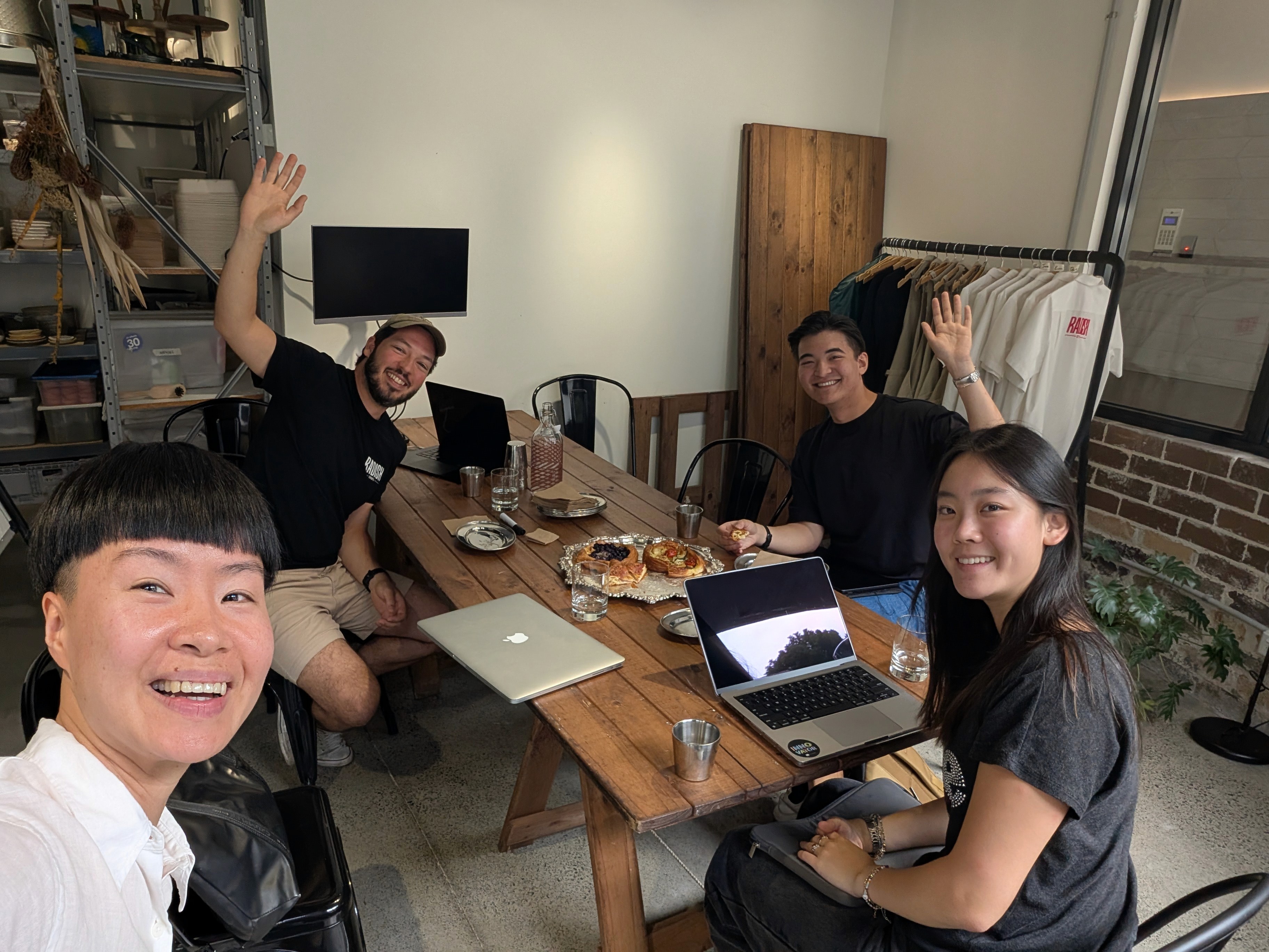 Four people around a long wooden table with laptops and pastries in a studio, two waving at the camera, a clothing rack behind them.