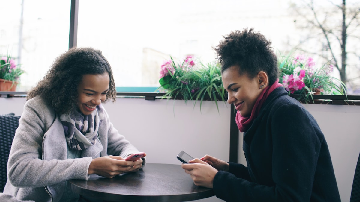 Two friends laughing over a phone at a cafe while comparing a group trip plan