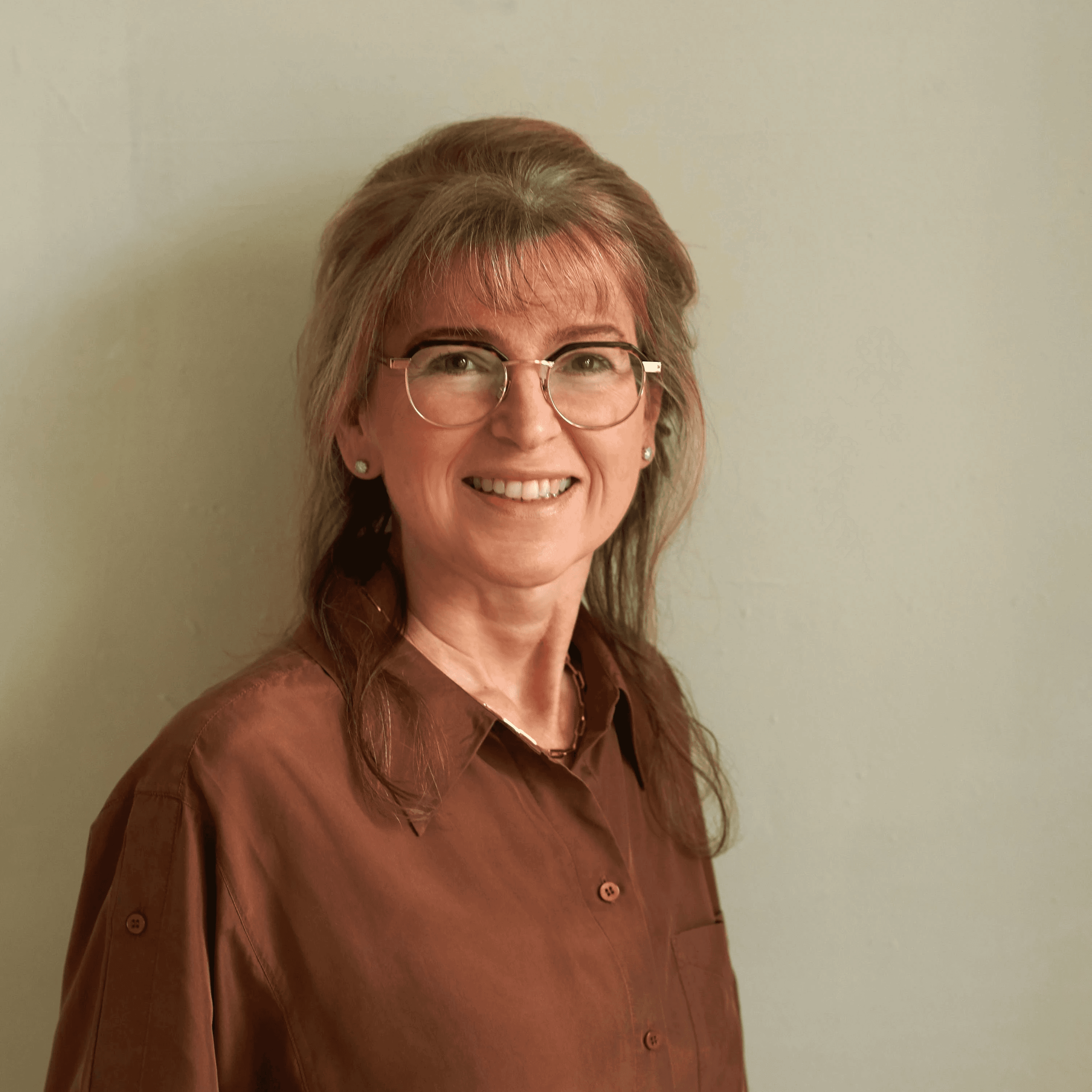 Portrait of Deborah Taylor Nash smiling, wearing a pair of black and gold framed glasses and a brown linen shirt, against a soft beige background.