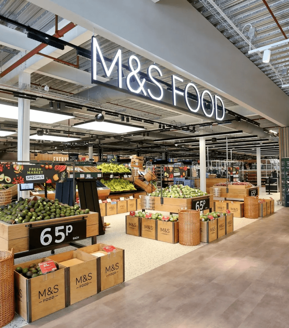 An M&S Food Hall interior with a prominent illuminated "M&S FOOD" sign, showcasing fresh produce displays. This image provides context for the in-store environment where the coffee machine product is located.