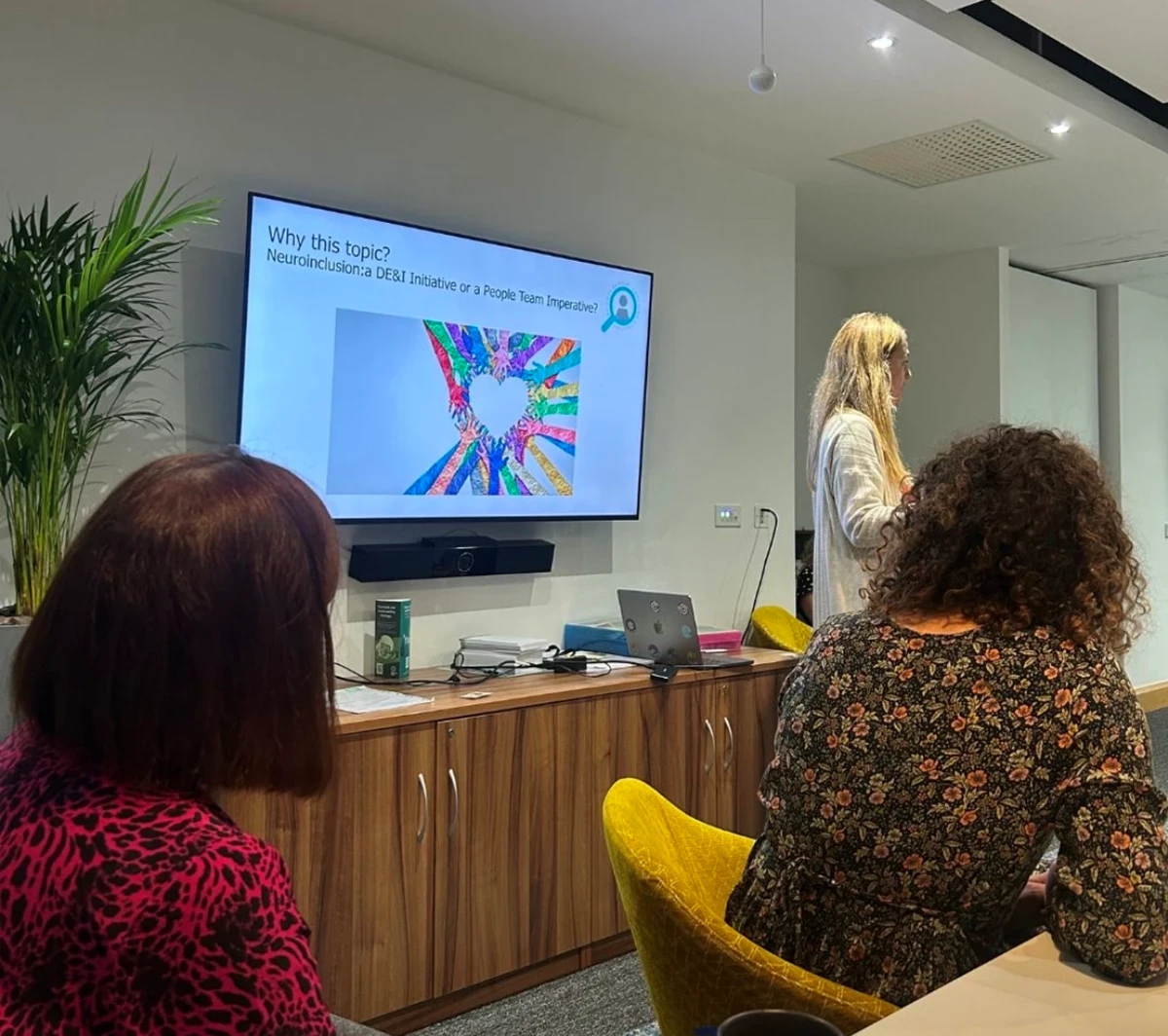 Person presenting in a meeting room with a screen showing “Why this topic?” and a colorful heart graphic, while two people sit at a table.