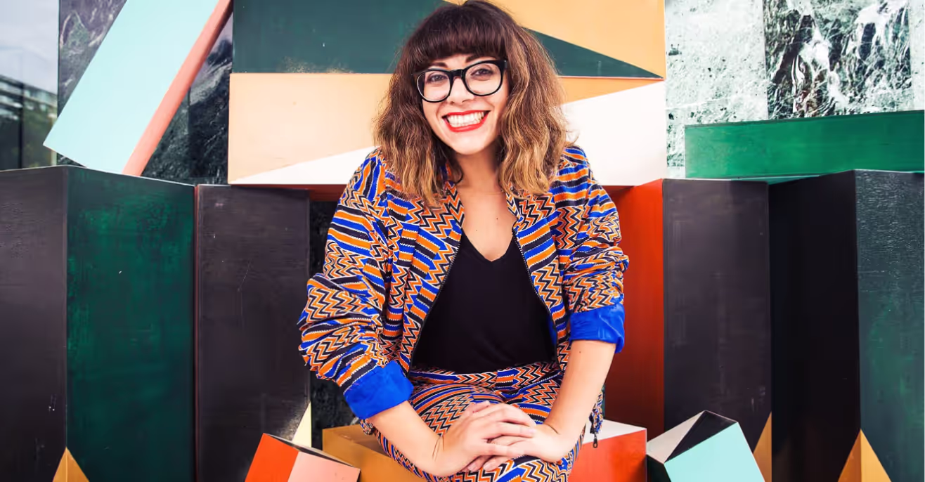Portrait of Jess Scully smiling in a brightly patterned jacket, standing among angular painted panels