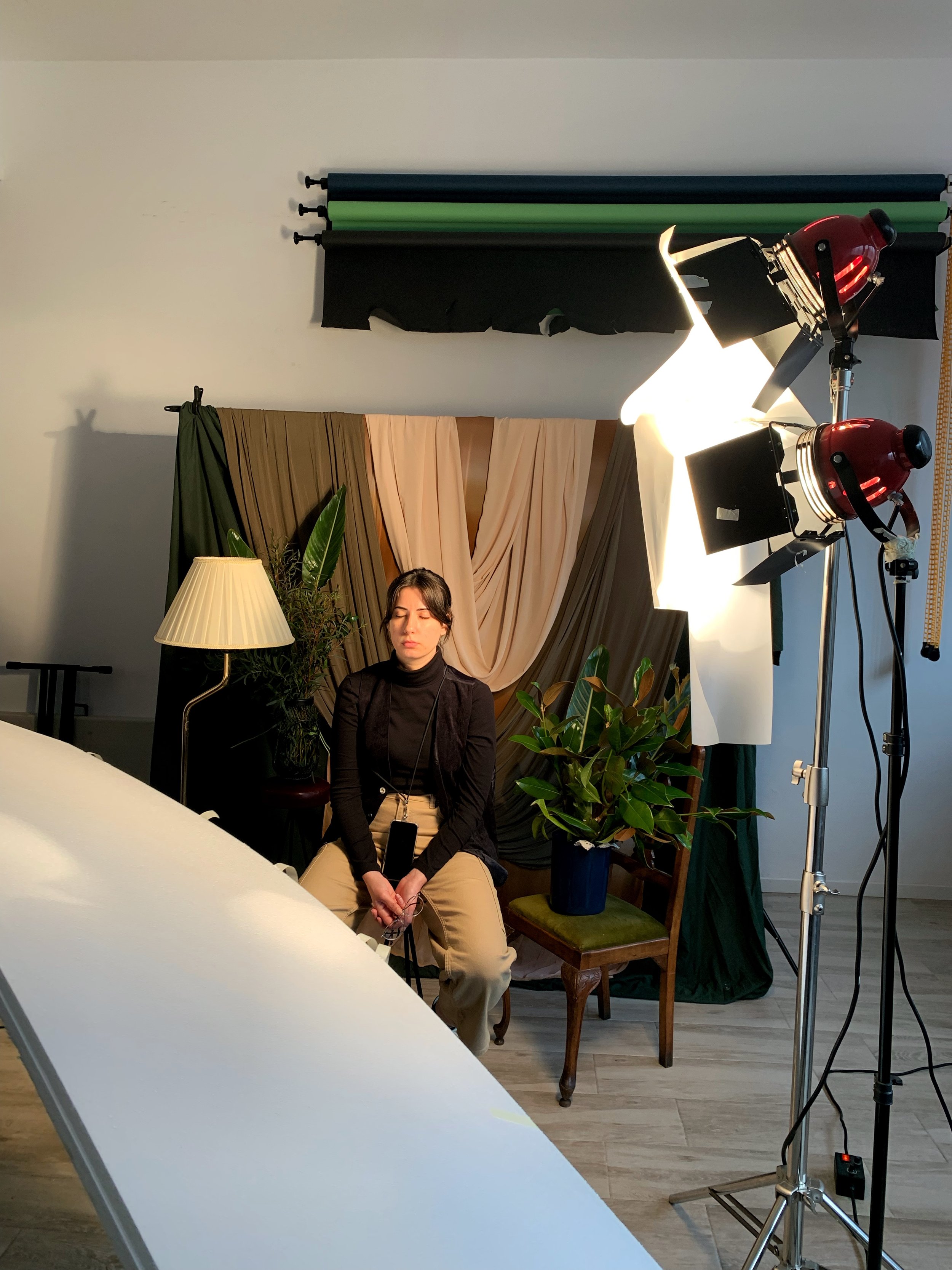 Woman sitting with eyes closed in a photography studio, surrounded by lighting equipment, plants, and curtains.