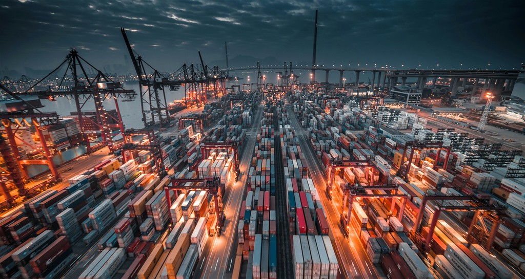 A large container terminal at night, illuminated by bright lights, with cranes and shipping containers creating a bustling industrial scene, representing global logistics and supply chain operations.