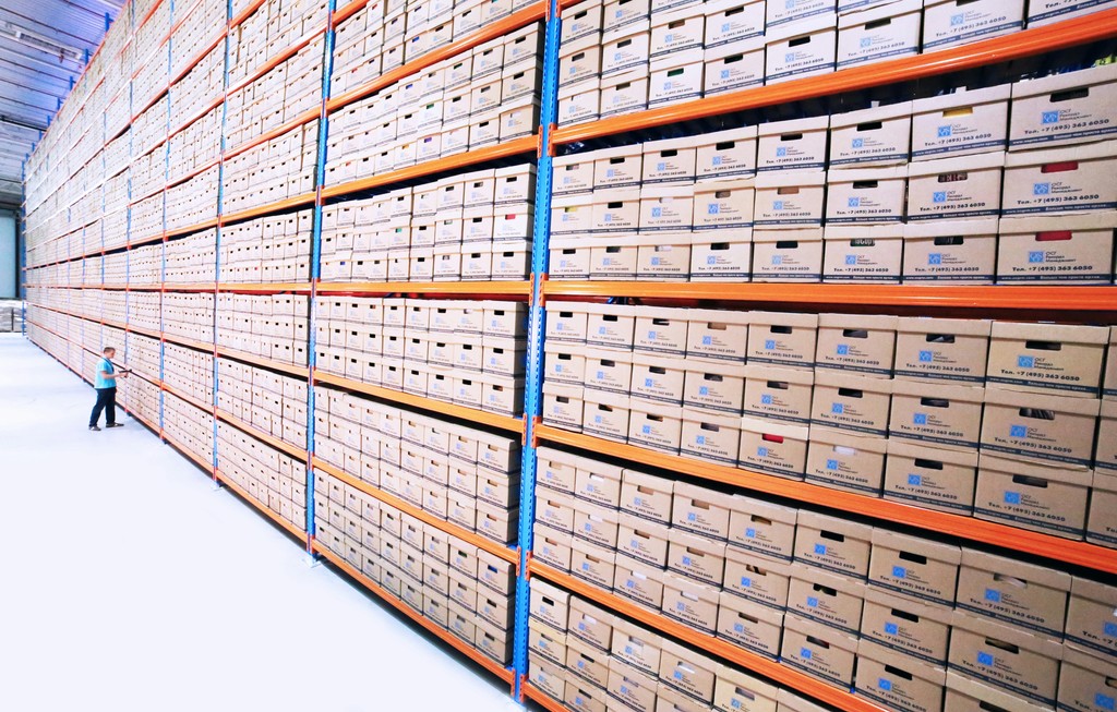 Large rows of high-stacked archive boxes in a warehouse with a person standing beside them, representing large-scale storage and data management.